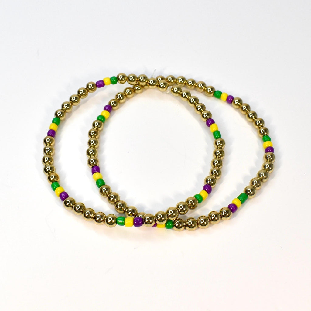 Two gold beaded bracelets with alternating green, yellow, and purple beads. The bracelets are circular and sit on a white surface. The gold beads are small and round.