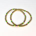 Two gold beaded bracelets with alternating green, yellow, and purple beads. The bracelets are circular and sit on a white surface. The gold beads are small and round.