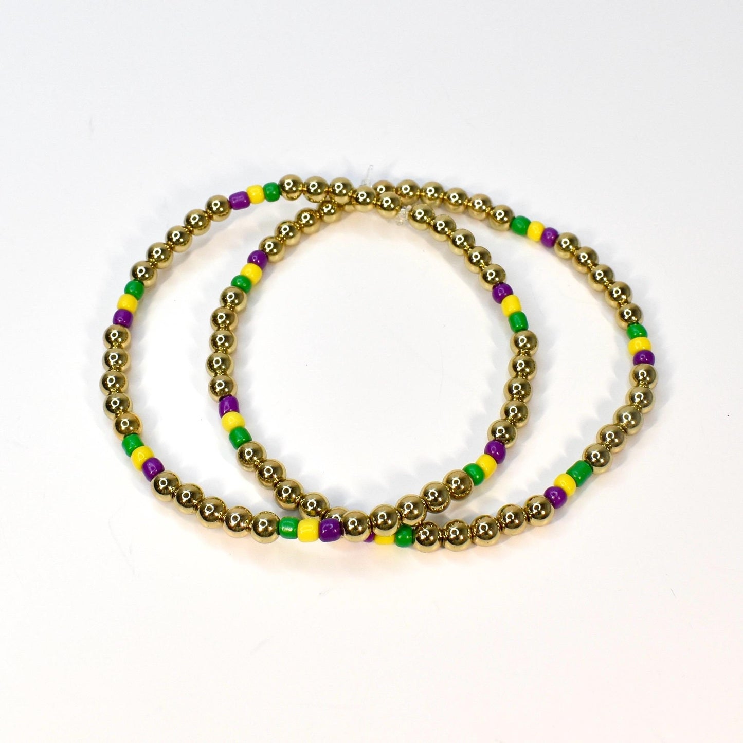 Two gold beaded bracelets with alternating green, yellow, and purple beads. The bracelets are circular and sit on a white surface. The gold beads are small and round.