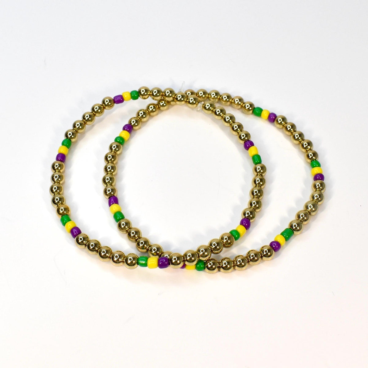 Two gold beaded bracelets with alternating green, yellow, and purple beads. The bracelets are circular and sit on a white surface. The gold beads are small and round.