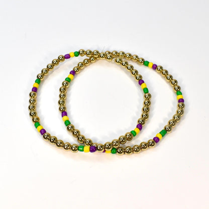 Two gold beaded bracelets with alternating green, yellow, and purple beads. The bracelets are circular and sit on a white surface. The gold beads are small and round.