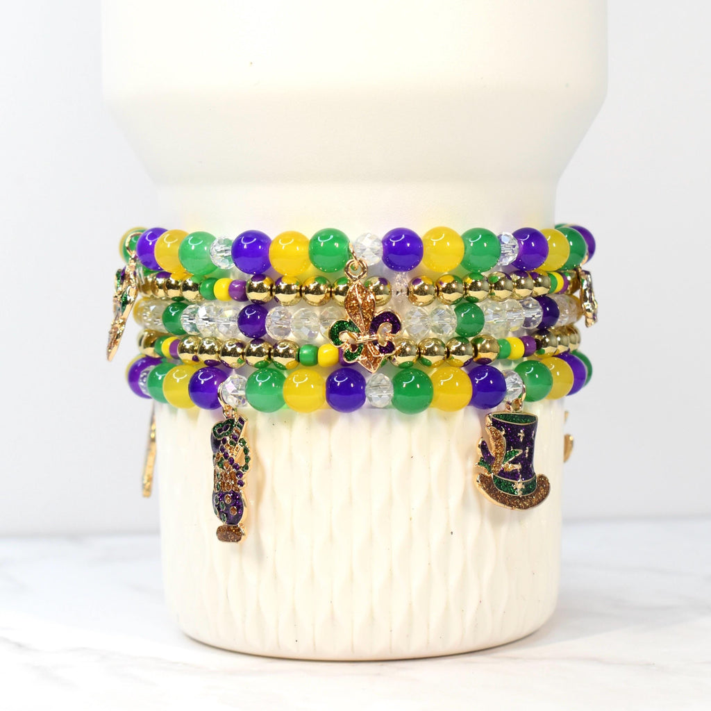 Stack of beaded bracelets in purple, green, yellow, and clear beads. Gold-colored charms include a fleur-de-lis, a top hat, and a decorative vase. The bracelets are stacked on a white ceramic base.