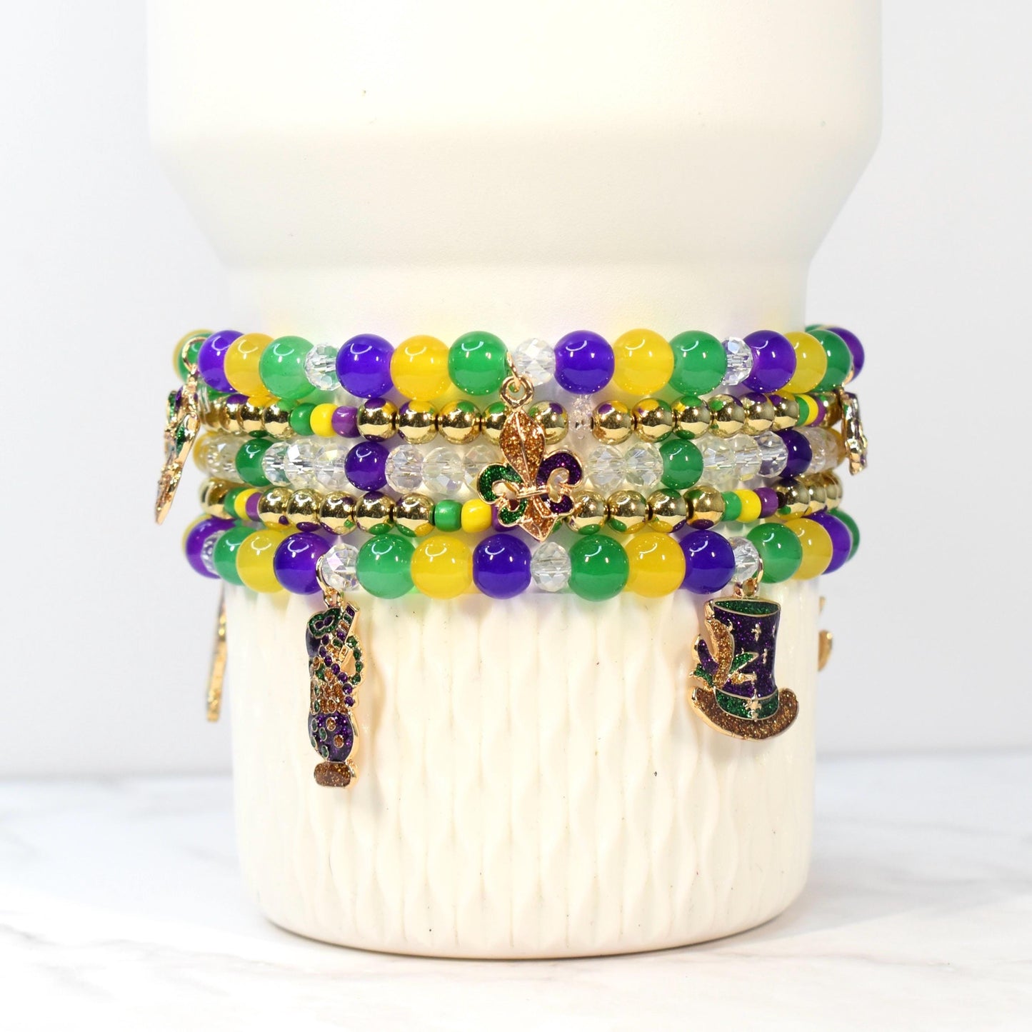 Stack of beaded bracelets in purple, green, yellow, and clear beads. Gold-colored charms include a fleur-de-lis, a top hat, and a decorative vase. The bracelets are stacked on a white ceramic base.