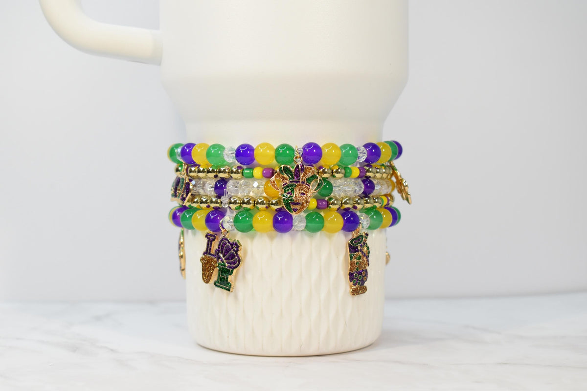 A stack of beaded bracelets in purple, green, yellow, and clear beads, adorned with charms. The bracelets are wrapped around a white mug. The charms include a fleur-de-lis and other Mardi Gras-themed designs.