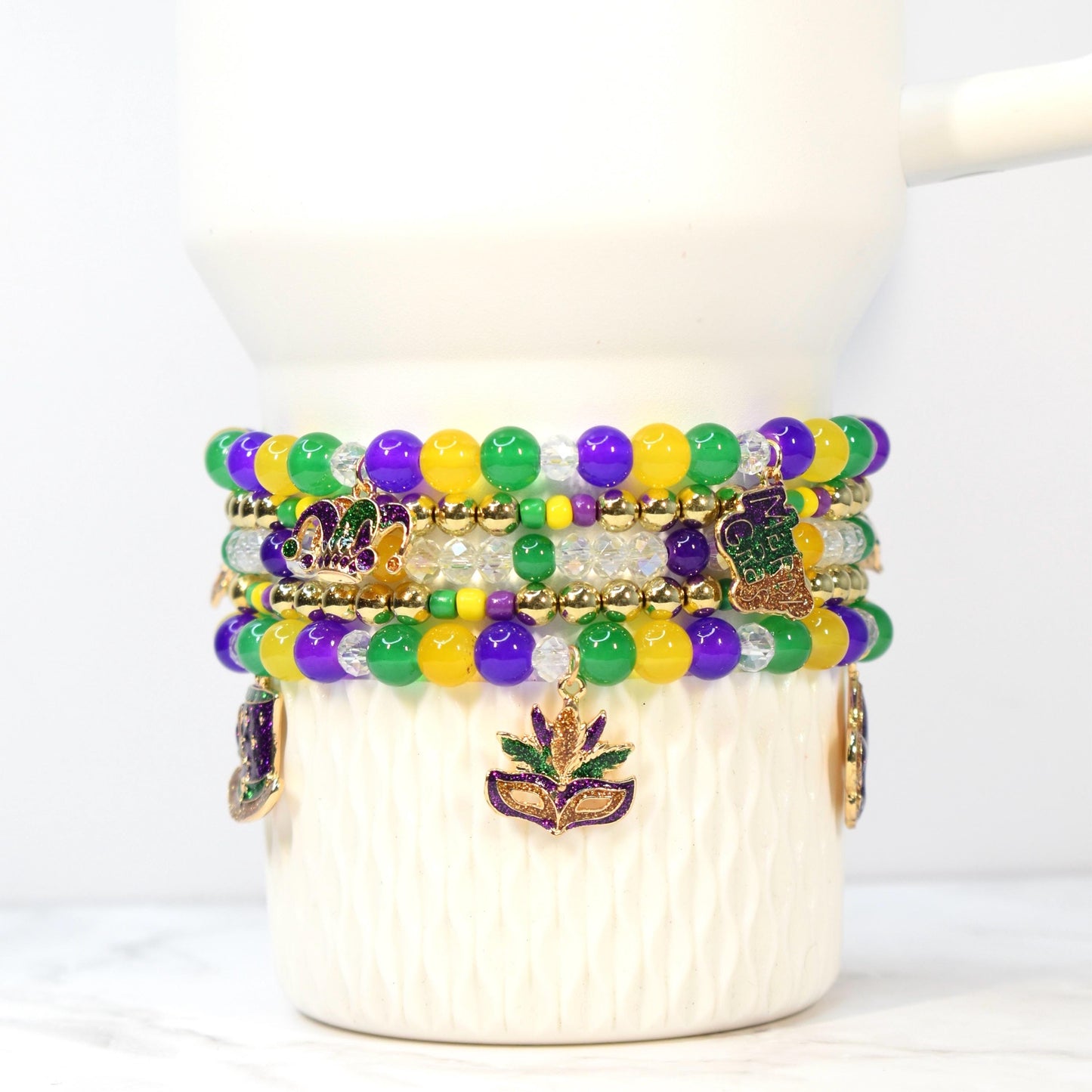Stack of beaded bracelets in Mardi Gras colors: purple, green, and yellow. Each bracelet features round beads and gold accents. Charms include a fleur-de-lis and a masquerade mask.