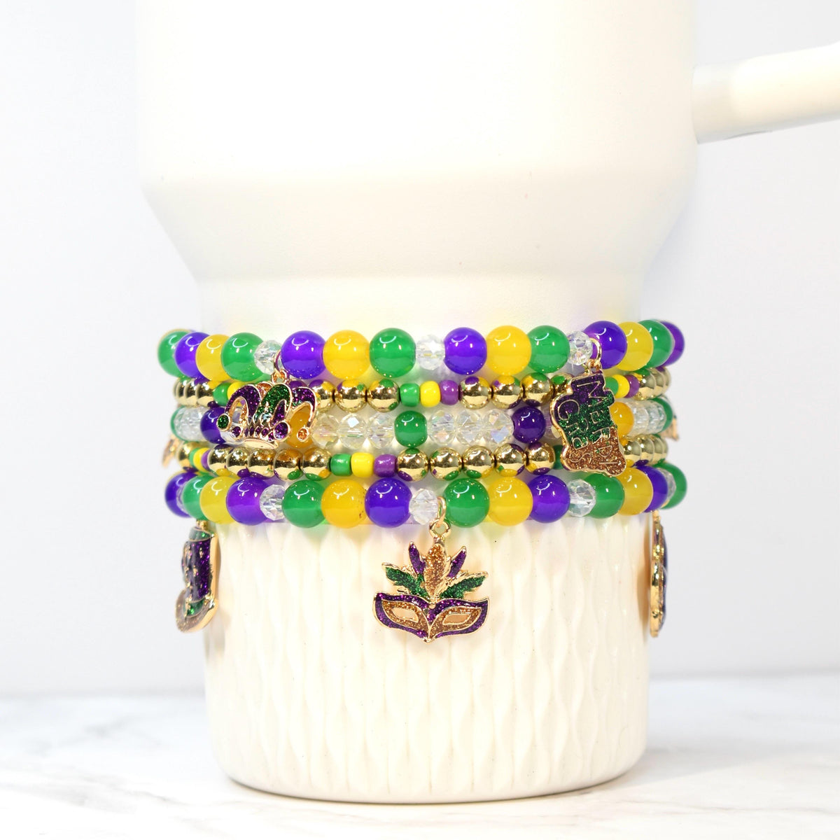 Stack of beaded bracelets in Mardi Gras colors: purple, green, and yellow. Each bracelet features round beads and gold accents. Charms include a fleur-de-lis and a masquerade mask.
