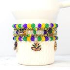 Stack of beaded bracelets in Mardi Gras colors: green, purple, and gold. Each bracelet features round beads and decorative charms, including a mask and a crown. The bracelets are displayed on a white ceramic cup.
