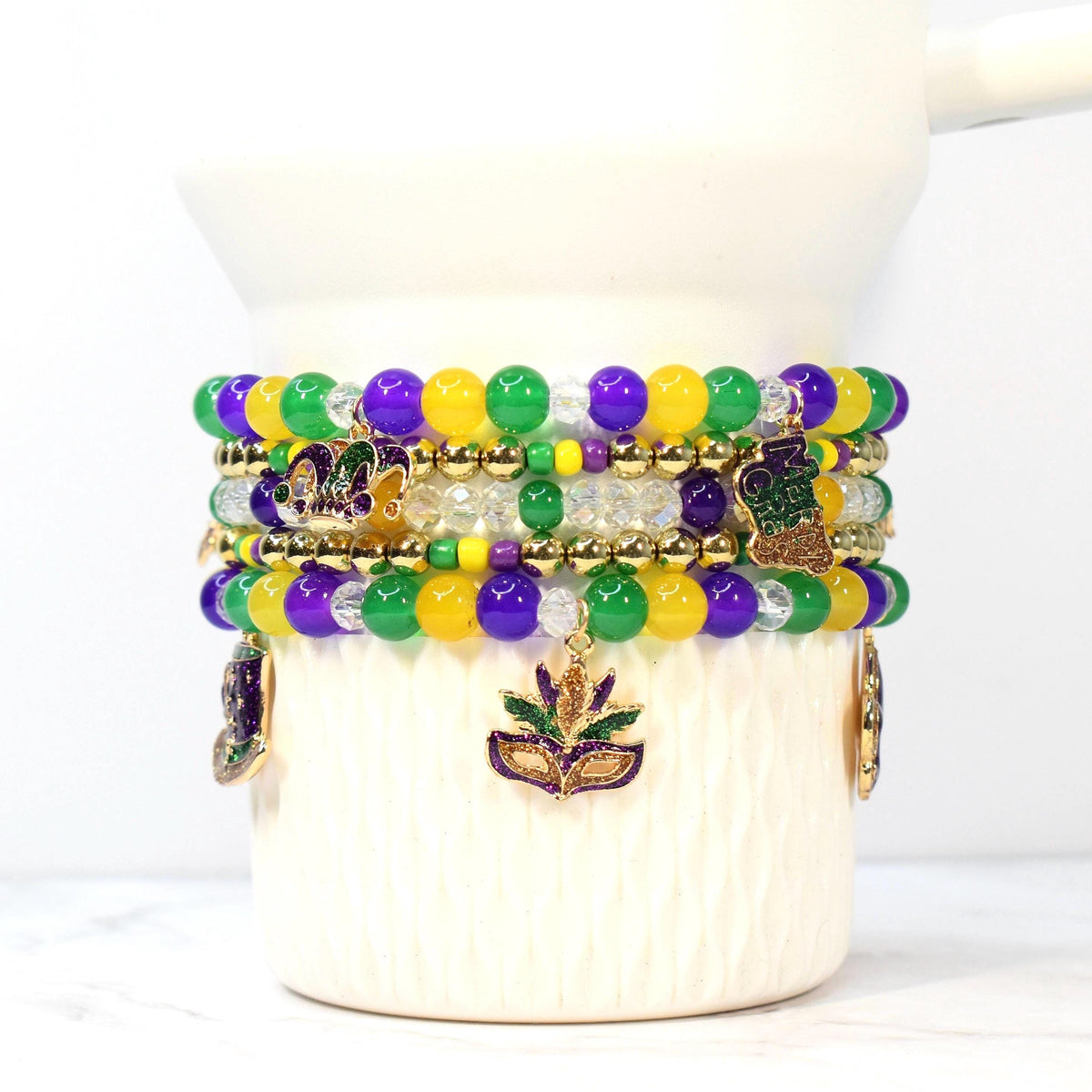 Stack of beaded bracelets in Mardi Gras colors: green, purple, and gold. Each bracelet features round beads and decorative charms, including a mask and a crown. The bracelets are displayed on a white ceramic cup.
