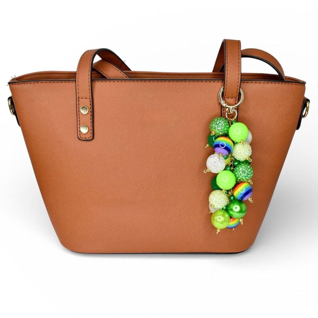 A brown tote bag with two handles and a decorative beaded charm. The charm features a variety of green, white, and rainbow-colored beads. The bag has a simple, clean design.