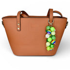 A brown tote bag with two handles and a decorative beaded charm. The charm features a variety of green, white, and rainbow-colored beads. The bag has a simple, clean design.