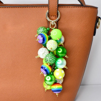 A colorful beaded keychain with a gold-tone clasp. The keychain features a cluster of vibrant, round beads in various colors, including green, yellow, blue, and rainbow patterns. The beads are attached to a gold-tone ring, designed to attach to a purse or bag.