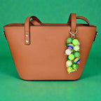 A brown tote bag with a beaded charm. The charm features a cluster of green, white, and rainbow-colored beads. The bag has two handles and gold-colored hardware.