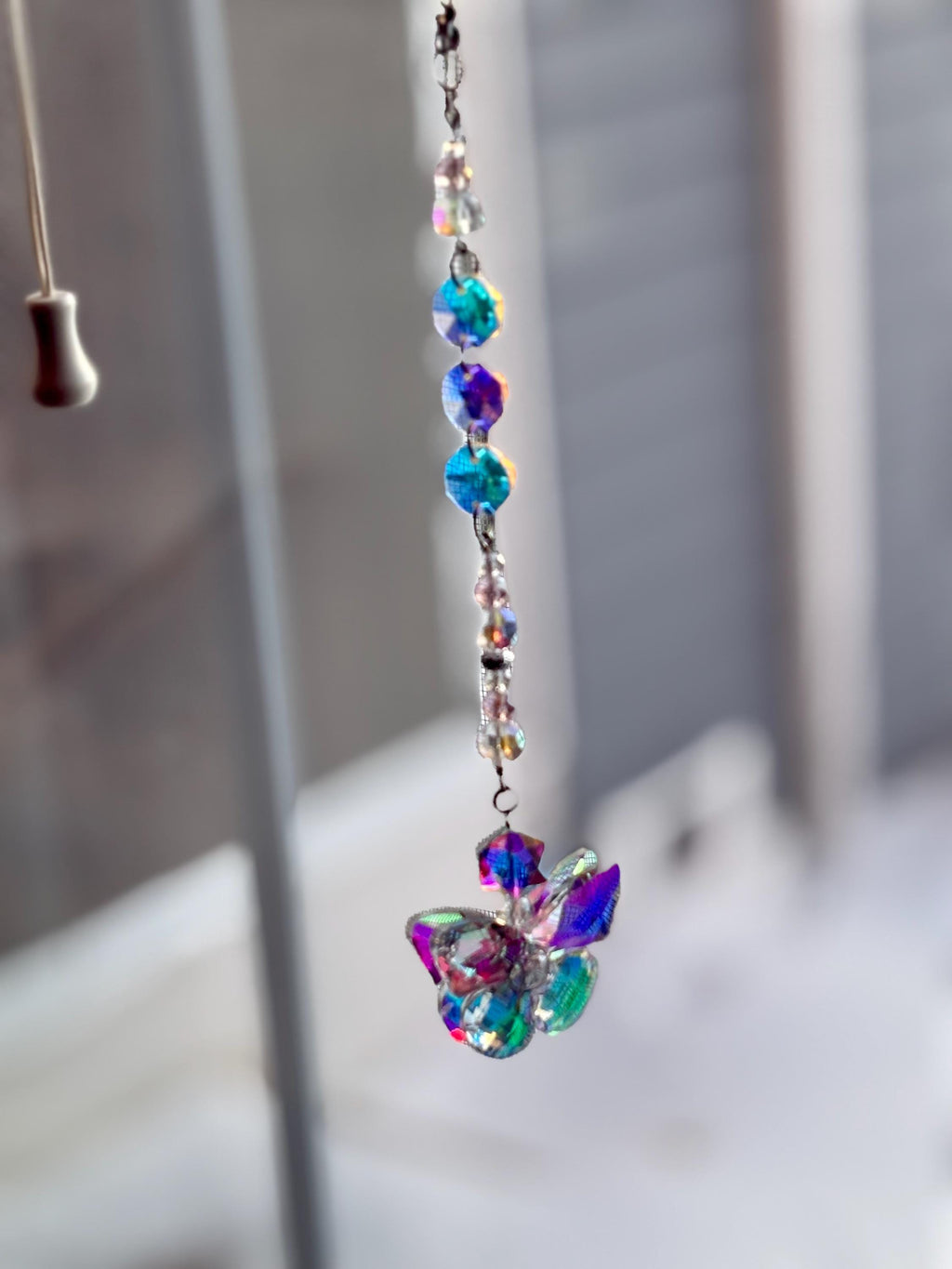 A hanging crystal suncatcher with a butterfly-shaped pendant. The suncatcher features iridescent, colorful crystals and beads strung on a silver chain. The butterfly pendant has a mix of blue, purple, and green hues.