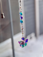 A hanging crystal suncatcher with a butterfly-shaped pendant. The suncatcher features iridescent, colorful crystals and beads strung on a silver chain. The butterfly pendant has a mix of blue, purple, and green hues.