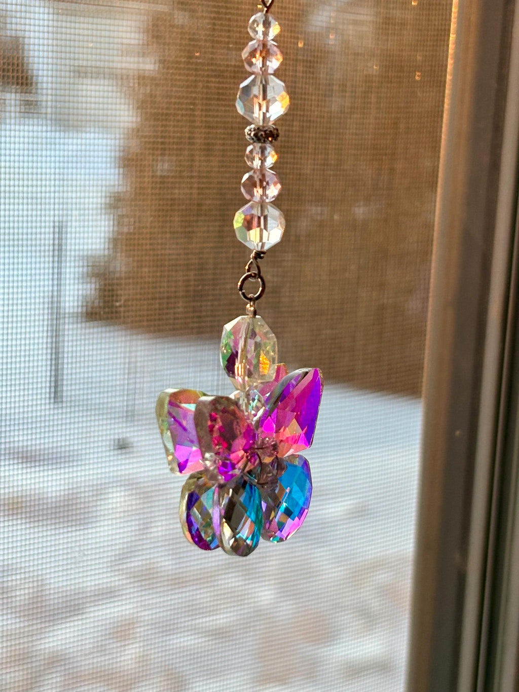 A hanging crystal suncatcher with a butterfly design. The butterfly is made of iridescent, faceted crystals in shades of pink, purple, and blue. The suncatcher is suspended by a string of clear, faceted beads.