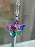 A decorative butterfly suncatcher with iridescent crystals. The butterfly has a faceted body and wings that reflect a spectrum of colors, including purple, blue, and green. It hangs from a string of clear and pink beads.