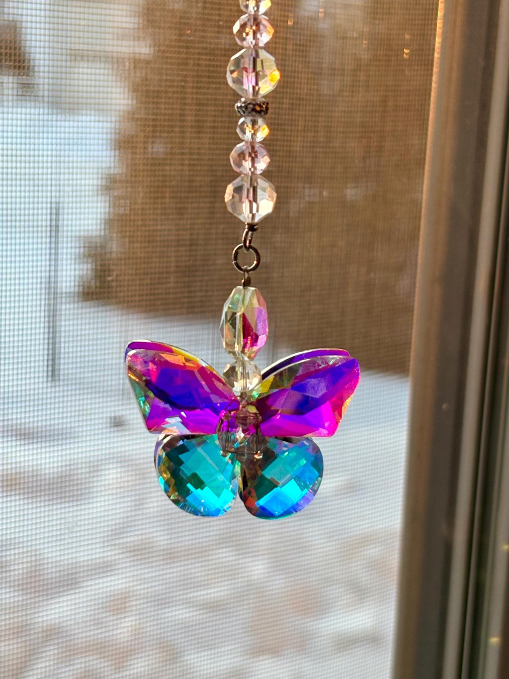 A colorful butterfly suncatcher with iridescent wings in shades of purple, pink, and teal. The butterfly is made of faceted glass and hangs from a strand of clear beads. The suncatcher is designed to catch the light and create a rainbow effect.