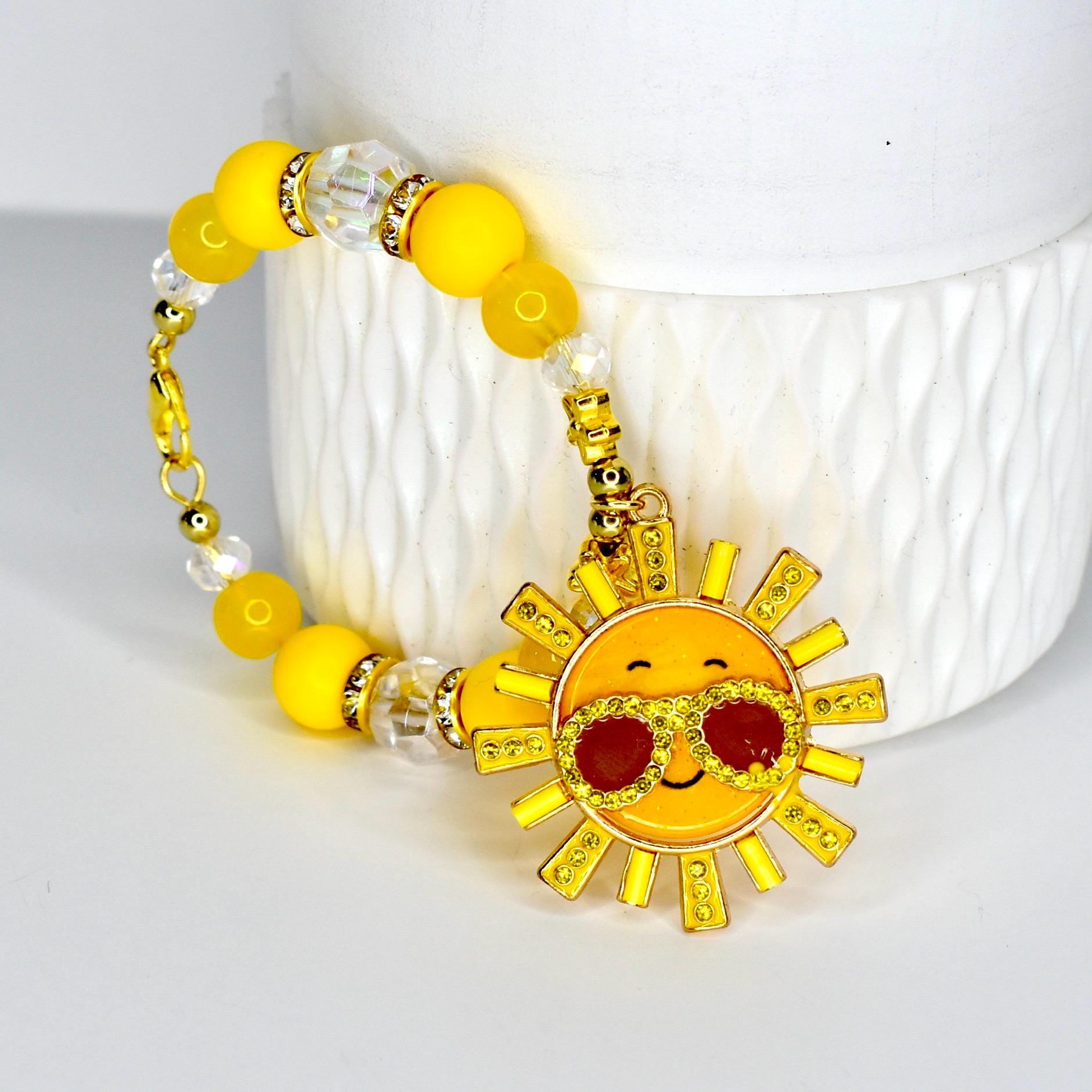 A yellow beaded handle bracelet with a gold clasp and a charm featuring a smiling sun wearing sunglasses. The bracelet has yellow and clear beads. The sun charm has red lenses and gold accents.