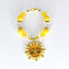 A yellow beaded bracelet with a gold clasp and a sun charm. The bracelet features yellow and clear beads, with a gold sun pendant wearing sunglasses. The sun charm has a smiling face and is surrounded by gold rays.
