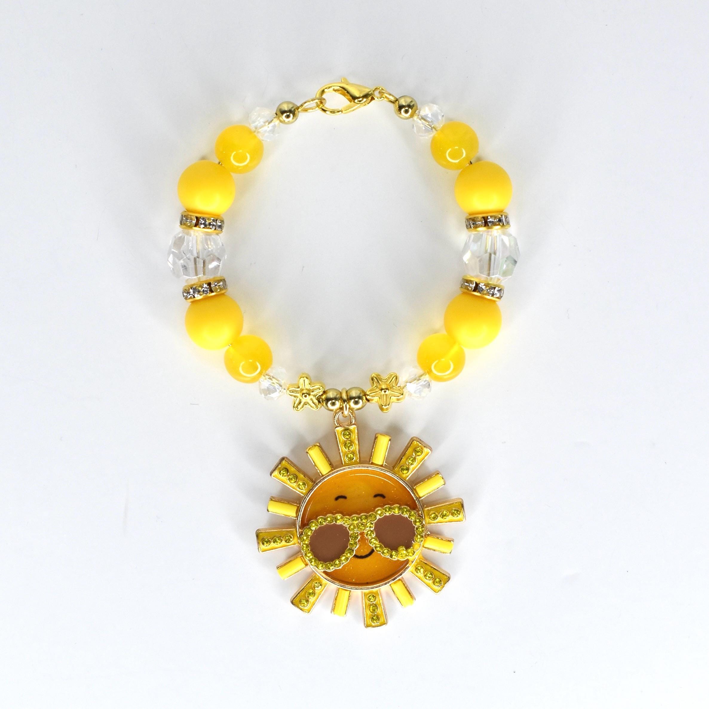 A yellow beaded bracelet with a gold clasp and a sun charm. The bracelet features yellow and clear beads, with a gold sun pendant wearing sunglasses. The sun charm has a smiling face and is surrounded by gold rays.