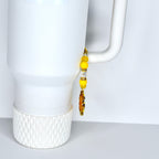 A white tumbler with a decorative yellow beaded charm on the handle. The charm features yellow beads, clear crystal beads, and a sunflower pendant. The tumbler has a white handle and a textured base.