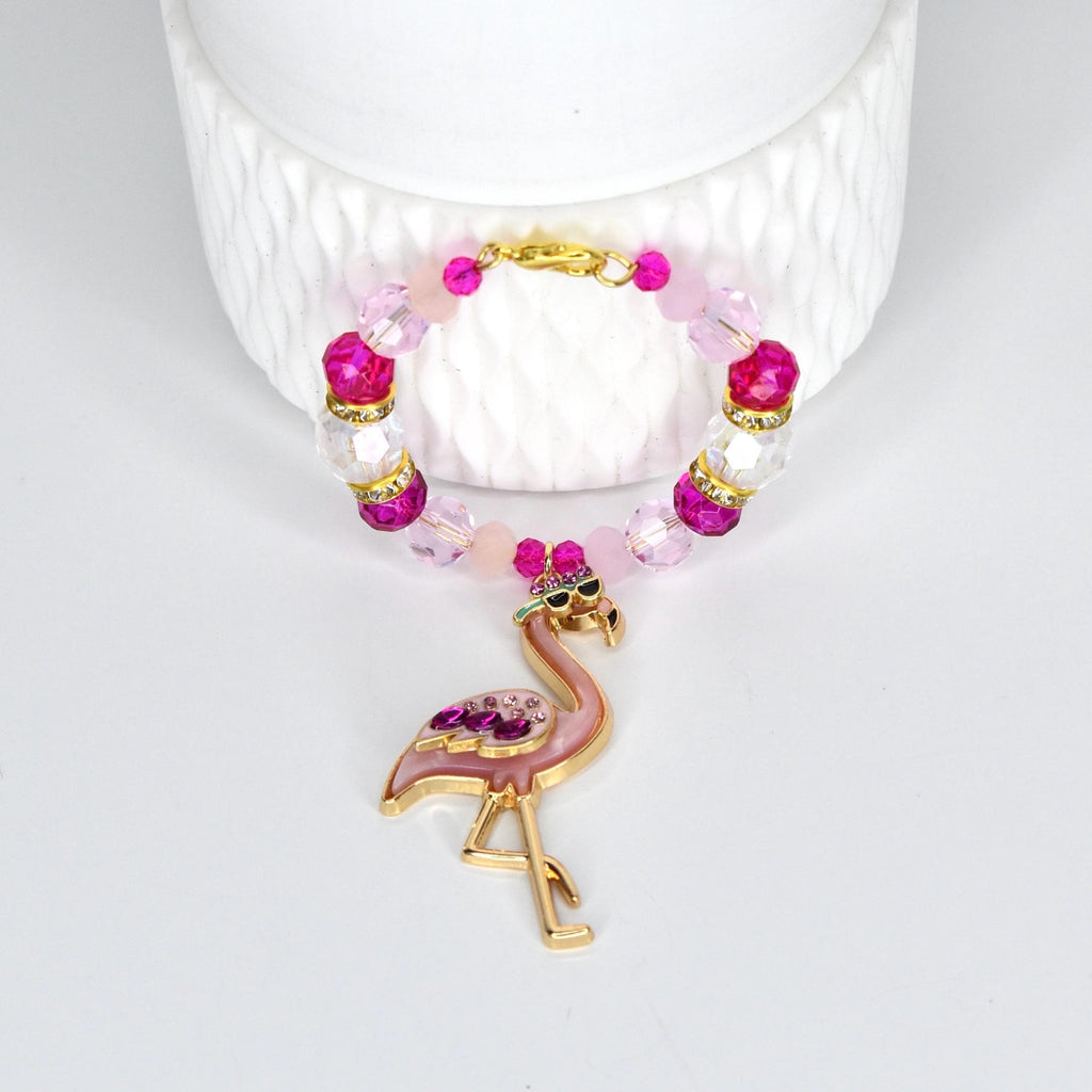 A pink and gold flamingo handle charm bracelet. The handle bracelet features a gold clasp and is adorned with pink, clear, and fuchsia beads. The flamingo charm is embellished with pink rhinestones and sunglasses.