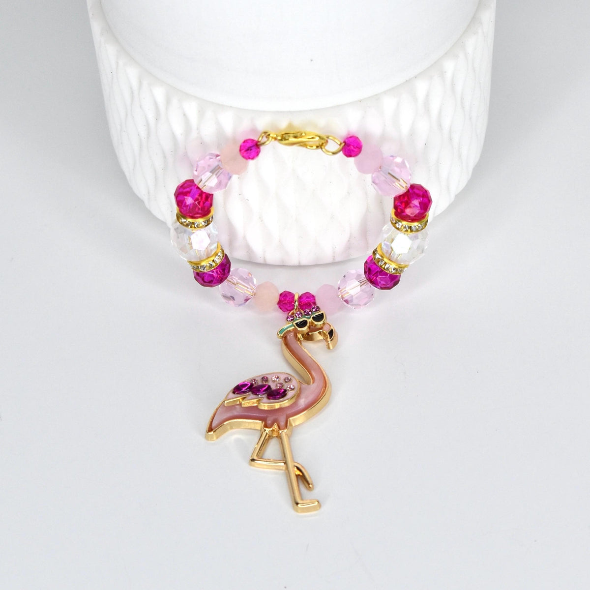 A pink and gold flamingo handle charm bracelet. The handle bracelet features a gold clasp and is adorned with pink, clear, and fuchsia beads. The flamingo charm is embellished with pink rhinestones and sunglasses.
