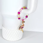 A beaded bracelet with a flamingo charm. The bracelet features pink, clear, and gold beads. The flamingo charm is gold with pink enamel and purple accents. The bracelet is hanging on a white object.