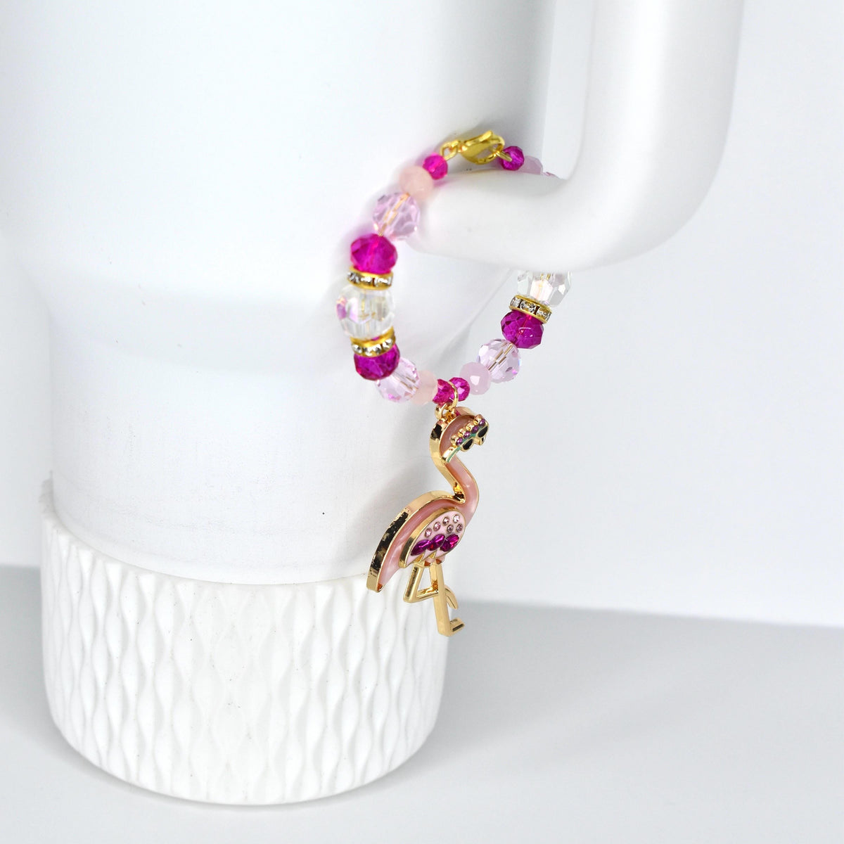A beaded bracelet with a flamingo charm. The bracelet features pink, clear, and gold beads. The flamingo charm is gold with pink enamel and purple accents. The bracelet is hanging on a white object.