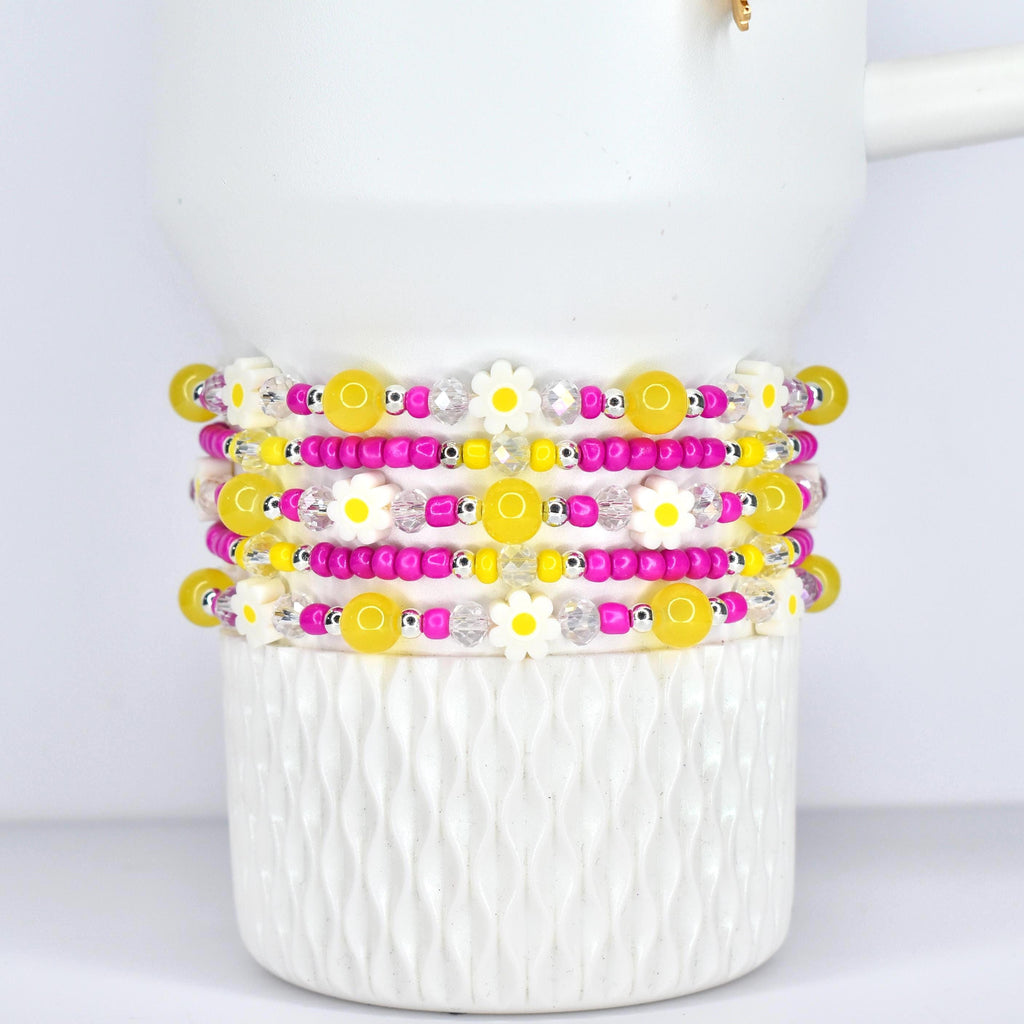 Stack of beaded bracelets with yellow, pink, and white daisy and bead accents. The bracelets are displayed on a white, textured ceramic cup. The beads are strung on elastic.
