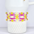 Stack of beaded bracelets with yellow, pink, and white daisy and bead accents. The bracelets are displayed on a white, textured ceramic cup. The beads are strung on elastic.