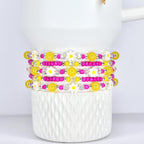 Stack of beaded bracelets with yellow, pink, and white daisy and bead accents. The bracelets are displayed on a white, textured ceramic cup. The beads are strung on elastic.
