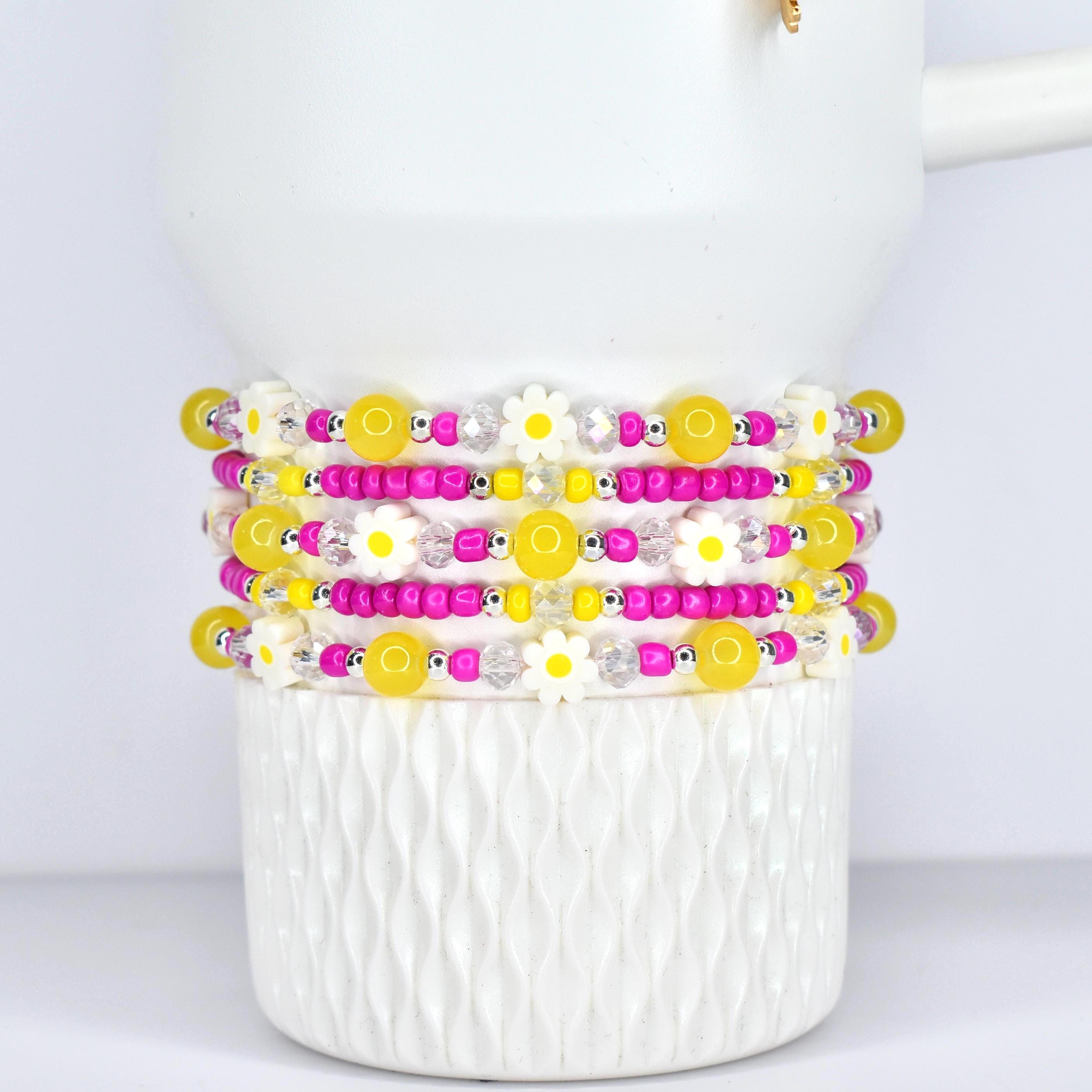 Stack of beaded bracelets with yellow, pink, and white daisy and bead accents. The bracelets are displayed on a white, textured ceramic cup. The beads are strung on elastic.