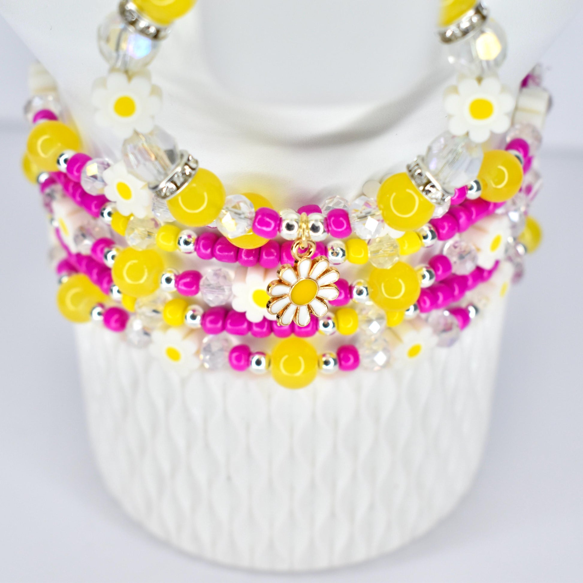 A multi-strand beaded bracelet stack featuring yellow, pink, and clear beads. The stack includes daisy-shaped charms and accents. The beads are strung together with silver-colored metal. The stack is displayed on a white form.