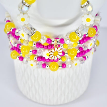 A multi-strand beaded bracelet stack featuring yellow, pink, and clear beads. The stack includes daisy-shaped charms and accents. The beads are strung together with silver-colored metal. The stack is displayed on a white form.