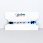 A beaded bracelet with the name "CASSIAN" spelled out in white letter beads. The bracelet features a mix of blue, black, and clear beads. The bracelet is displayed on a white card with the "Calathea Collective" logo.