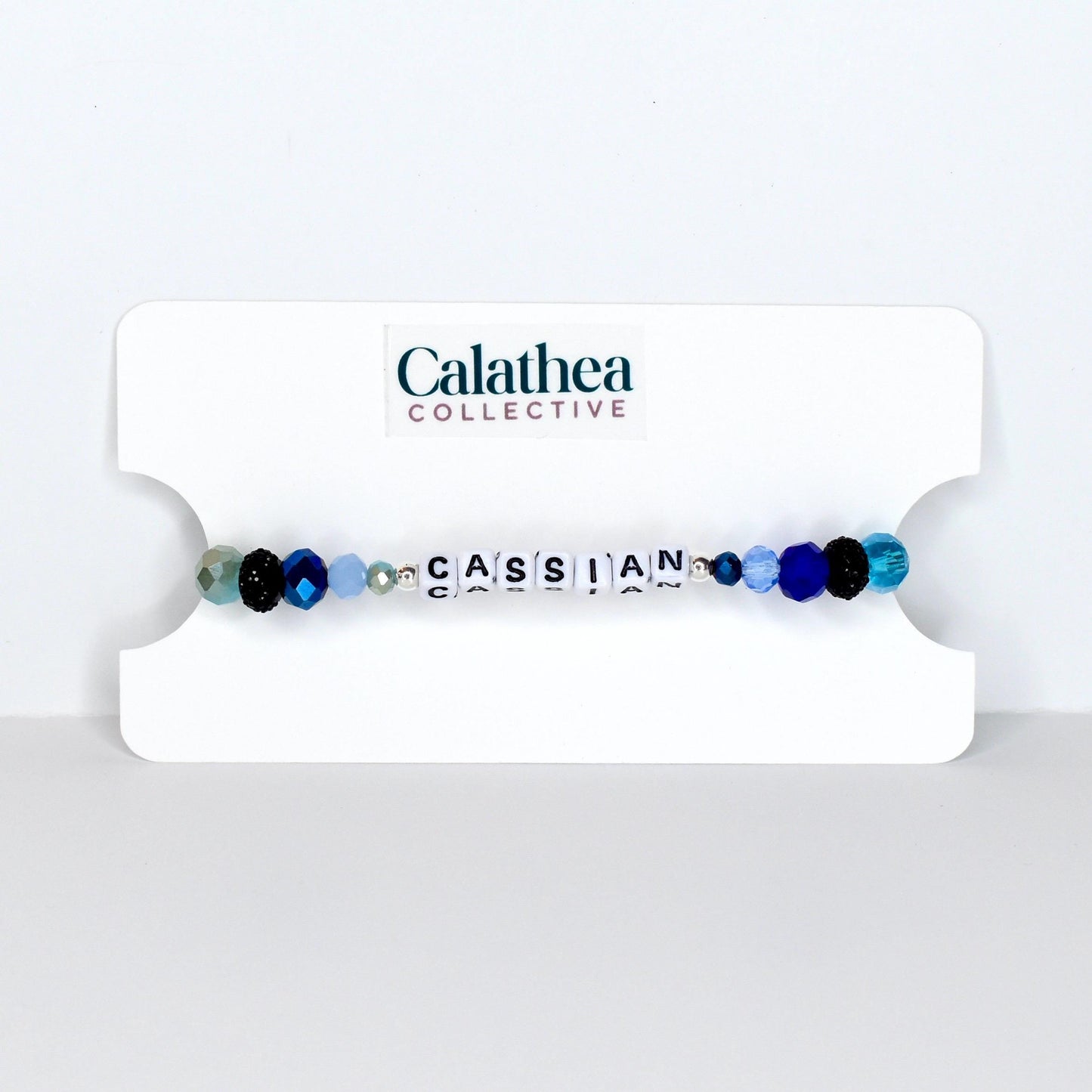 A beaded bracelet with the name "CASSIAN" spelled out in white letter beads. The bracelet features a mix of blue, black, and clear beads. The bracelet is displayed on a white card with the "Calathea Collective" logo.