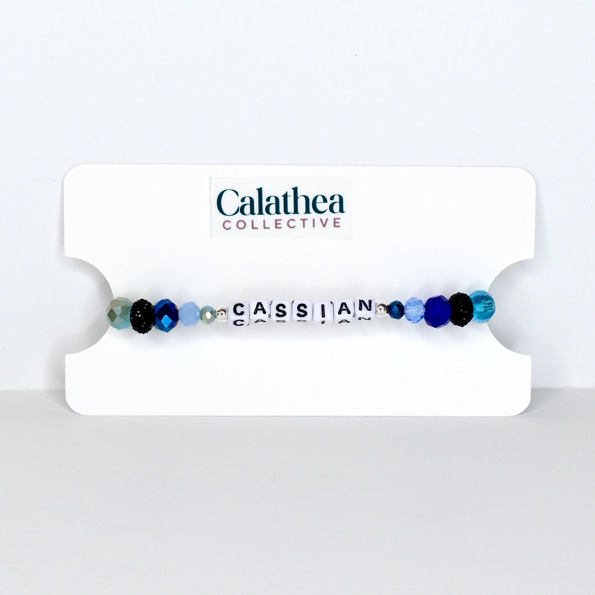 A beaded bracelet with the name "CASSIAN" spelled out in white letter beads. The bracelet features a mix of blue, black, and clear beads. The bracelet is displayed on a white card with the "Calathea Collective" logo.