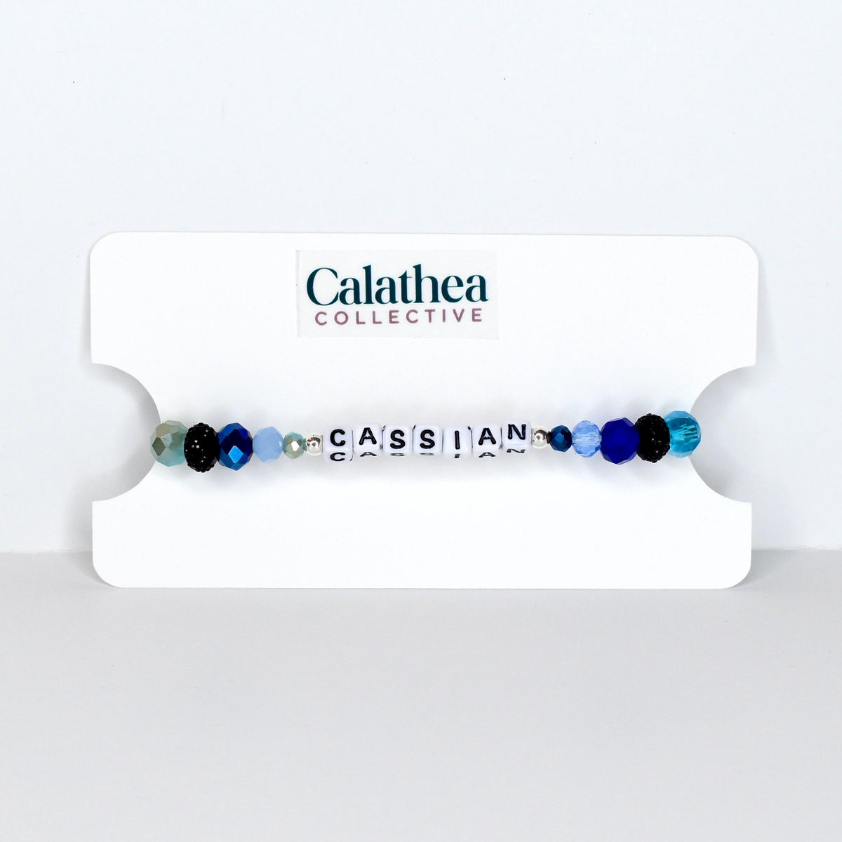 A beaded bracelet with the name "CASSIAN" spelled out in white letter beads. The bracelet features a mix of blue, black, and clear beads. The bracelet is displayed on a white card with the "Calathea Collective" logo.