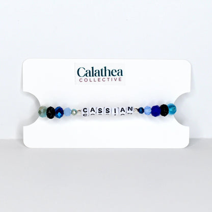 A beaded bracelet with the name "CASSIAN" spelled out in white letter beads. The bracelet features a mix of blue, black, and clear beads. The bracelet is displayed on a white card with the "Calathea Collective" logo.