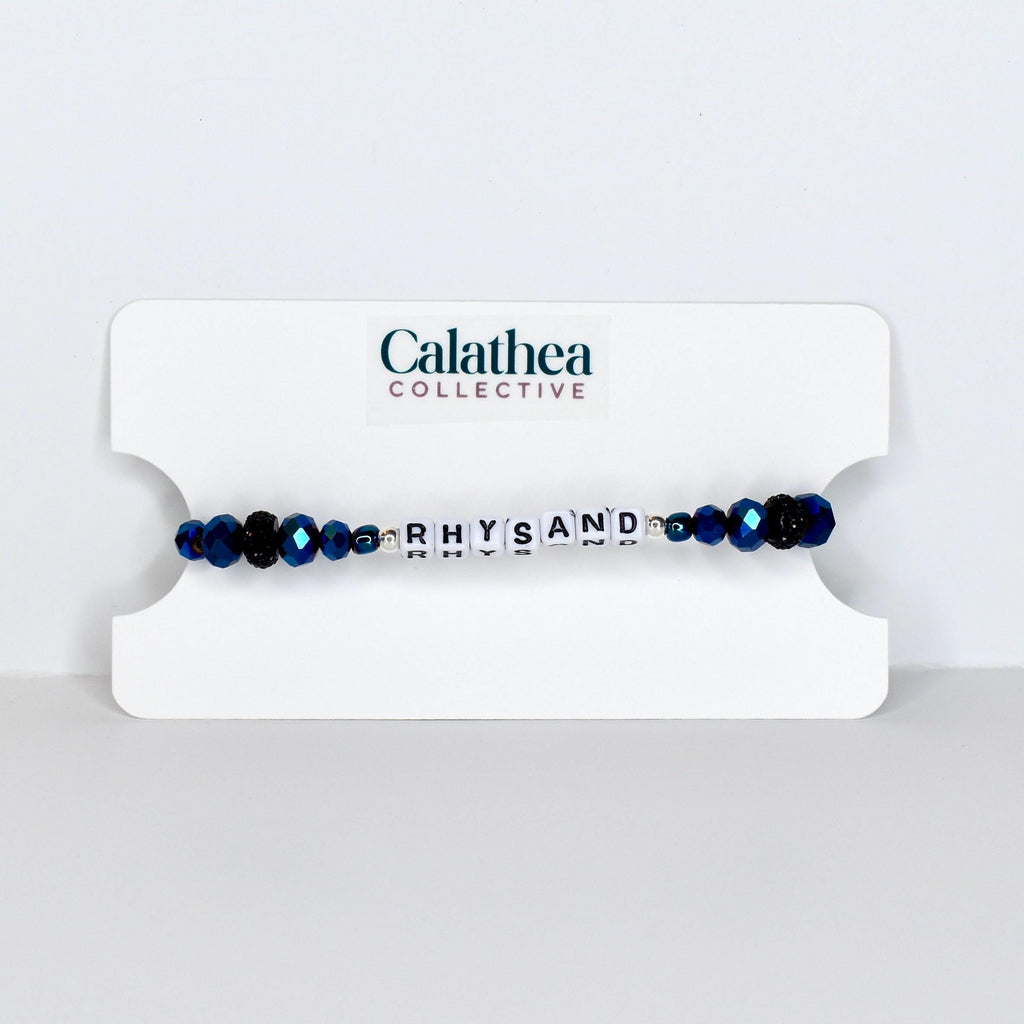 A beaded bracelet with the name "RHYSAND" spelled out in white letter beads. The bracelet features dark blue faceted beads and is displayed on a white card with the "Calathea Collective" logo.