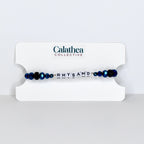 A beaded bracelet with the name "RHYSAND" spelled out in white letter beads. The bracelet features dark blue faceted beads and is displayed on a white card with the "Calathea Collective" logo.