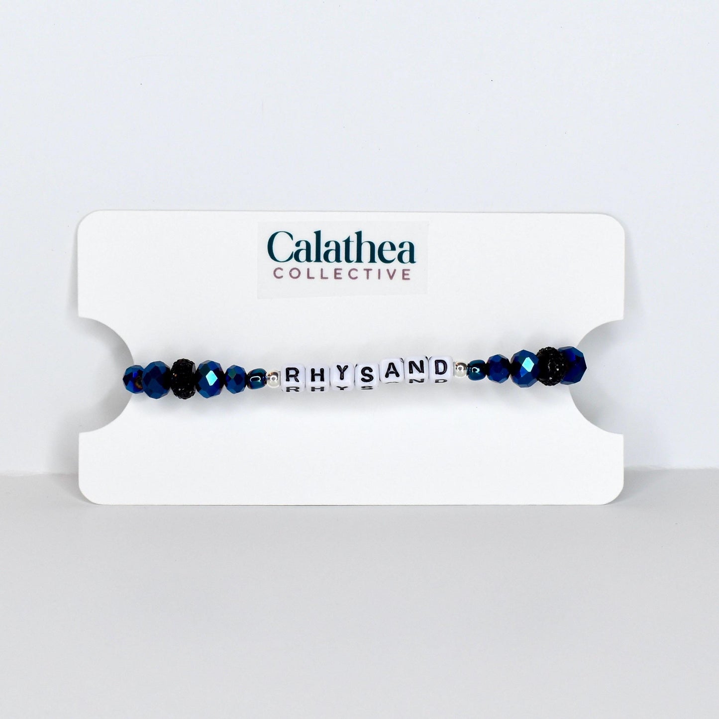 A beaded bracelet with the name "RHYSAND" spelled out in white letter beads. The bracelet features dark blue faceted beads and is displayed on a white card with the "Calathea Collective" logo.
