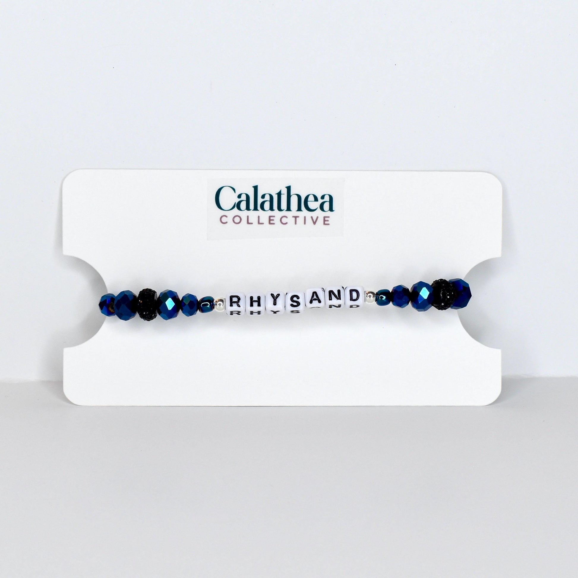 A beaded bracelet with the name "RHYSAND" spelled out in white letter beads. The bracelet features dark blue faceted beads and is displayed on a white card with the "Calathea Collective" logo.