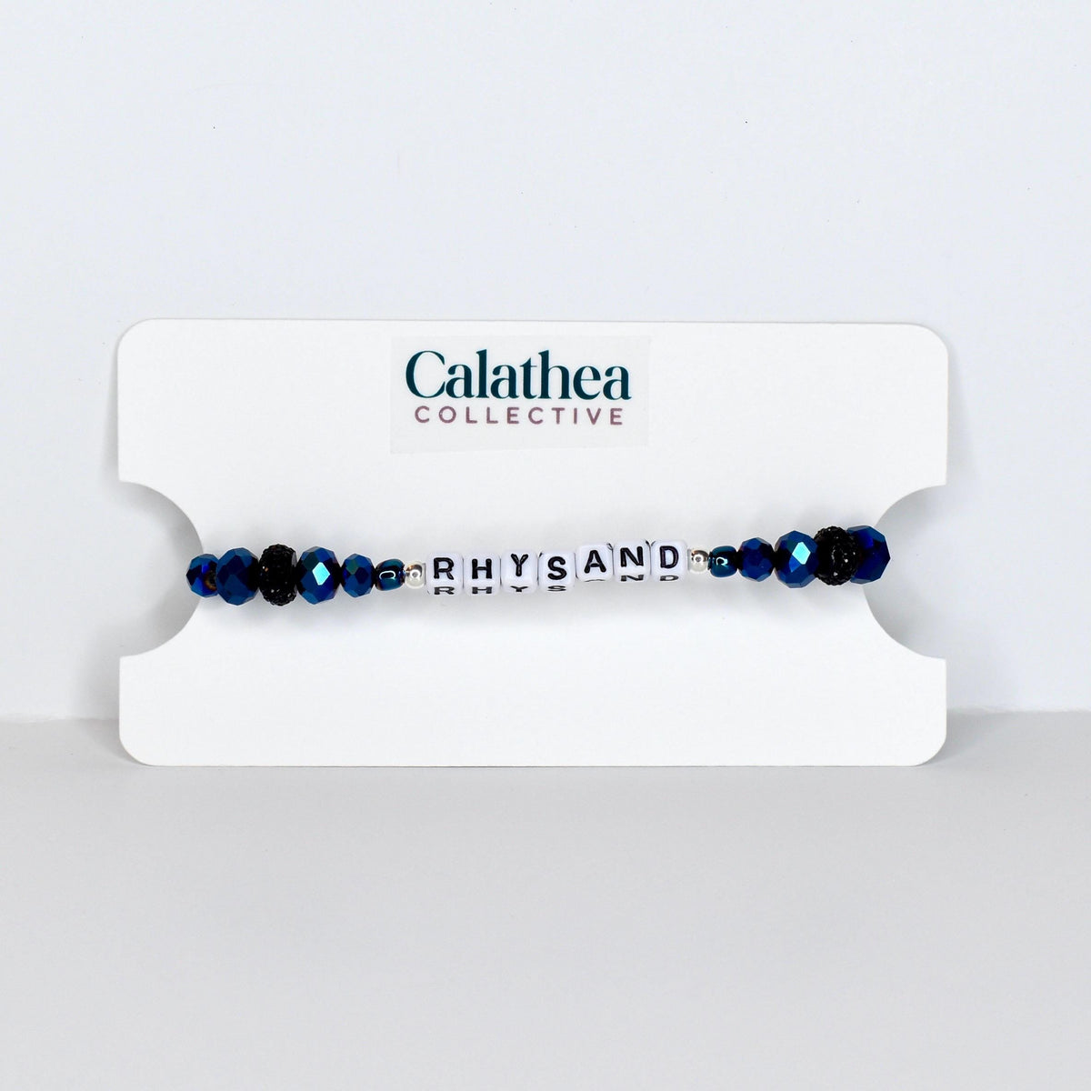 A beaded bracelet with the name "RHYSAND" spelled out in white letter beads. The bracelet features dark blue faceted beads and is displayed on a white card with the "Calathea Collective" logo.