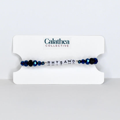 A beaded bracelet with the name "RHYSAND" spelled out in white letter beads. The bracelet features dark blue faceted beads and is displayed on a white card with the "Calathea Collective" logo.