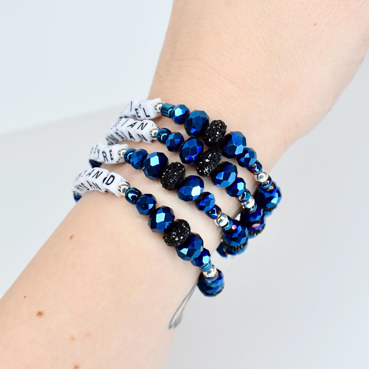 A set of four wrap bracelets. The bracelets feature faceted blue beads, black beaded accents, and white bands with black text. The bracelets are wrapped around a wrist.