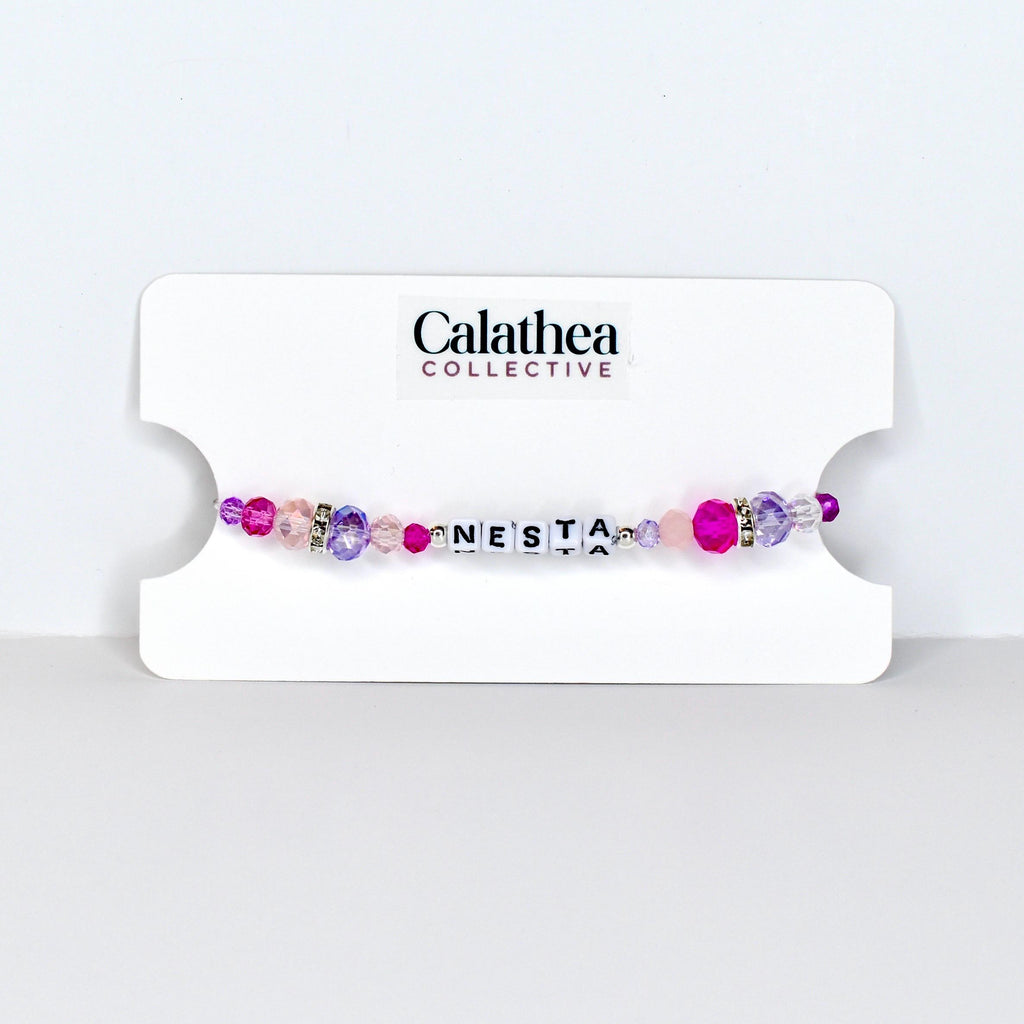 A beaded bracelet with the name "NESTA" spelled out in white letter beads. The bracelet features a mix of pink, purple, and clear beads, with a few silver accent beads. The bracelet is displayed on a white card with the "Calathea Collective" logo.