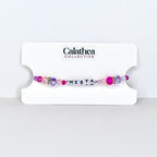 A beaded bracelet with the name "NESTA" spelled out in white letter beads. The bracelet features a mix of pink, purple, and clear beads, with a few silver accent beads. The bracelet is displayed on a white card with the "Calathea Collective" logo.