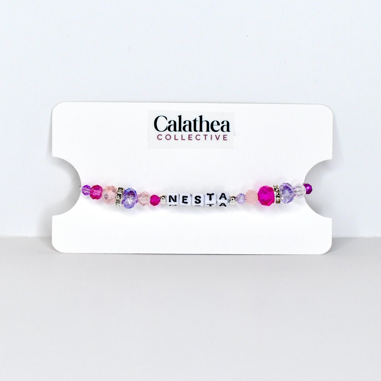 A beaded bracelet with the name "NESTA" spelled out in white letter beads. The bracelet features a mix of pink, purple, and clear beads, with a few silver accent beads. The bracelet is displayed on a white card with the "Calathea Collective" logo.